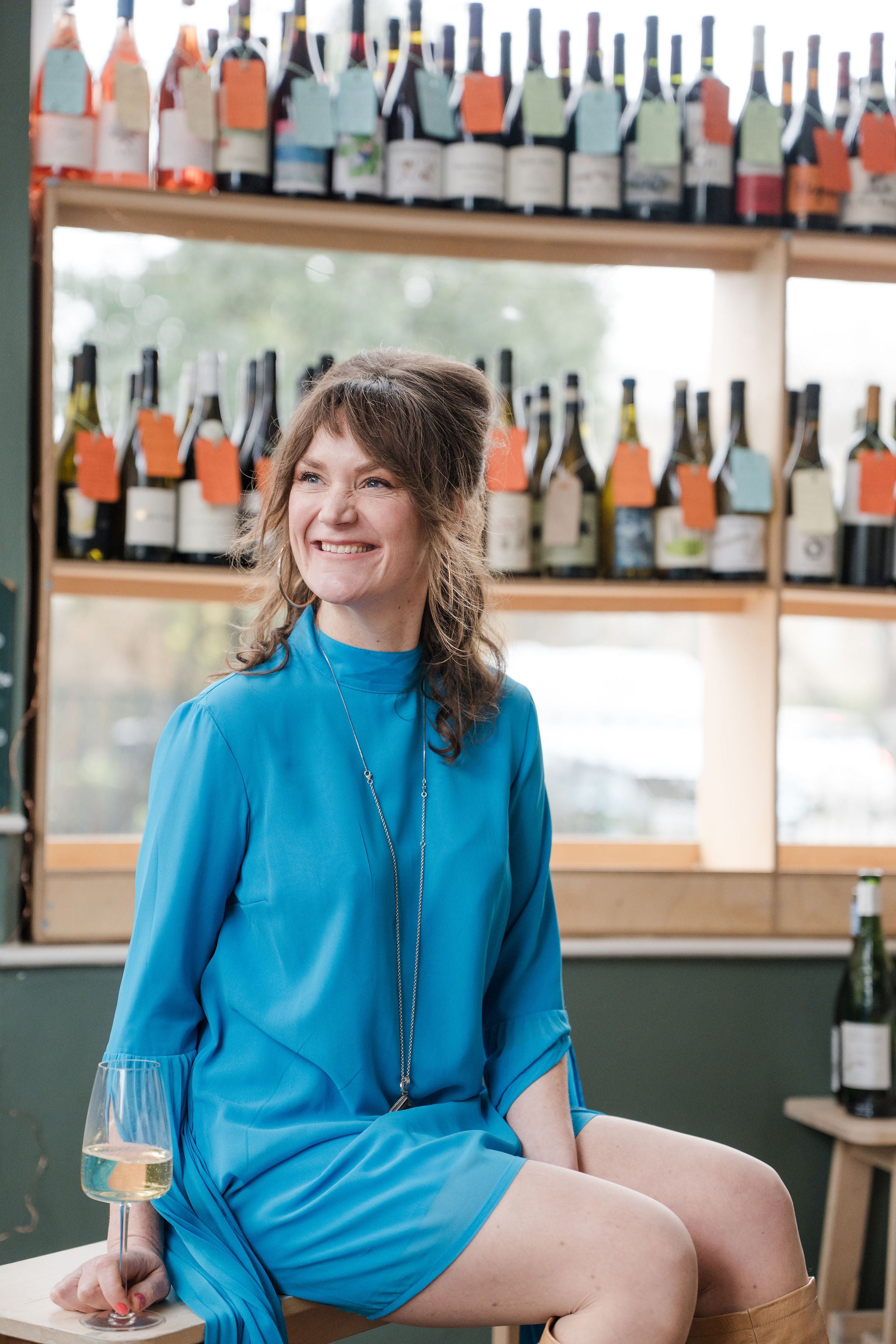 Sophia Luckett in a blue dress sitting in a wine cellar with wine bottles on shelves behind her.