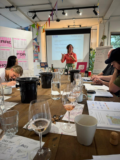 Group of people at a table with wine glasses and papers, presenter at front with screen